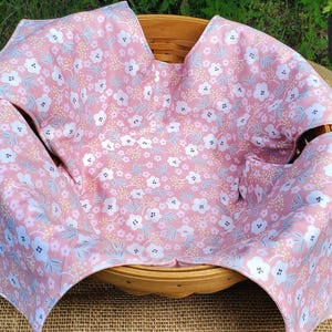 May include: A round wooden basket lined with a pink fabric featuring a repeating pattern of white flowers with dark centers, light blue leaves, and small yellow dots. The fabric drapes over the edges of the basket, which has a woven texture.