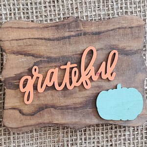 May include: A wooden sign with a distressed brown finish and a carved, scalloped edge. The sign features the word "grateful" in orange lettering and a light blue pumpkin cutout.