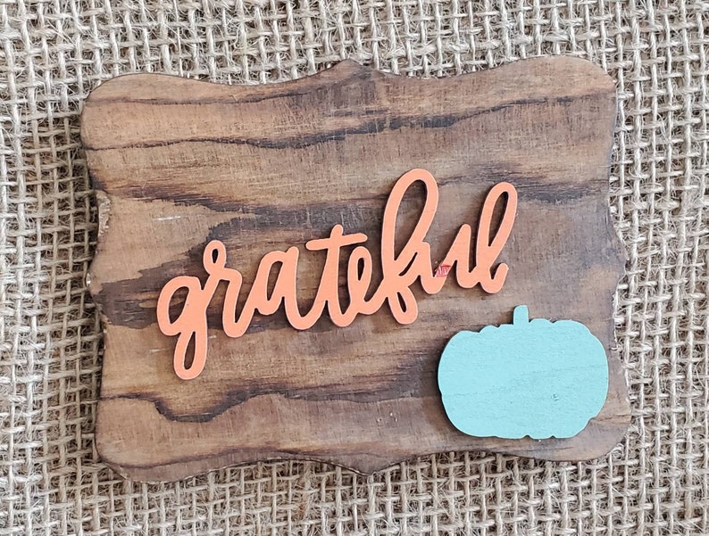 May include: A wooden sign with a distressed brown finish and a carved, scalloped edge. The sign features the word "grateful" in orange lettering and a light blue pumpkin cutout.