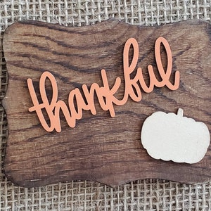 May include: A wooden sign with the word "thankful" in orange letters and a white pumpkin cutout. The sign has a rustic wood grain finish.