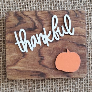 May include: A wooden sign with the word "thankful" written in white letters. A small orange pumpkin is in the bottom right corner of the sign.