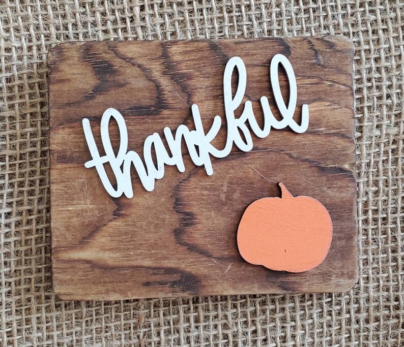 May include: A wooden sign with the word "thankful" written in white letters. A small orange pumpkin is in the bottom right corner of the sign.