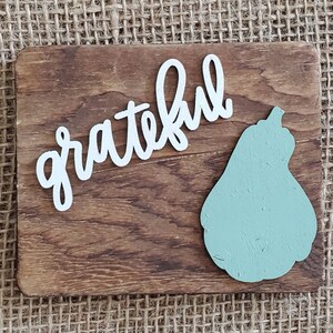 May include: A wooden sign with the word "grateful" written in white cursive lettering. A light green pear-shaped cutout is on the right side of the sign.