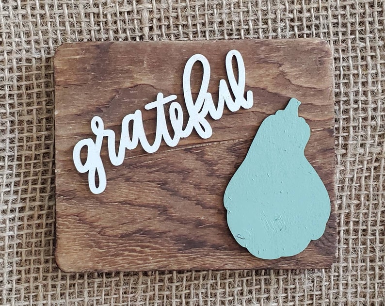 May include: A wooden sign with the word "grateful" written in white cursive lettering. A light green pear-shaped cutout is on the right side of the sign.