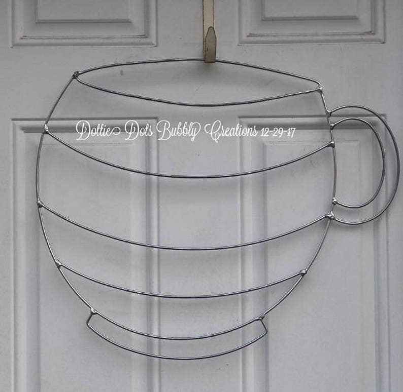 May include: A silver metal wire coffee cup wreath with a handle. The wreath is made of multiple layers of wire and is shaped like a coffee cup.