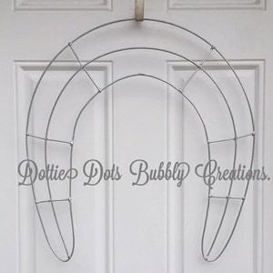 May include: A silver metal wreath frame with a horseshoe shape and a wire grid pattern. The frame is hanging on a white door.