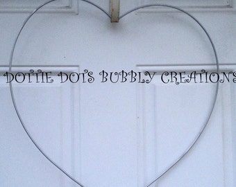 Könnte beinhalten: Eine silberne, herzförmige Kranzform aus Metall, die an einem Haken an einer weißen Tür hängt. Der Text "Dottie Dot's Bubbly Creations" ist unterhalb des Kranzes sichtbar.