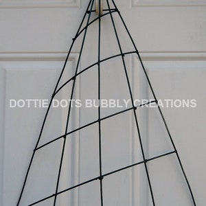 May include: A black wire frame in the shape of a cone, hanging from a hook on a white door. The frame is made of wire that is twisted together at the intersections. The frame is a craft supply for making wreaths or other decorations.