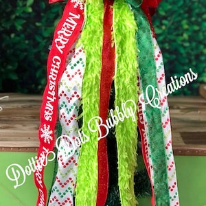 May include: A Christmas tree topper with a red, white, and green ribbon bow. The ribbon has the words "Merry Christmas" and "Merry Xmas" printed on it. The topper also includes green and red tinsel and a green fluffy ribbon.
