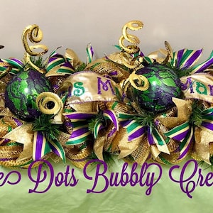 May include: A Mardi Gras themed centerpiece with gold, green, and purple ribbon, mesh, and ornaments. The centerpiece features the words "Mardi Gras" and "Miss Mardi Gras".