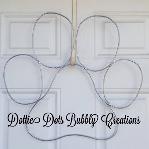 May include: A silver metal paw print wreath hanging on a white door. The wreath is made of wire and has a loop for hanging. The text "Dottie Dots Bubbly Creations" is written across the bottom of the wreath.