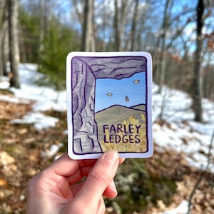 May include: A rectangular sticker featuring a stylized illustration of a rock formation with the text "FARLEY LEDGES." The design includes a blue sky, falling leaves, and a landscape. The sticker is held against a blurred forest background.