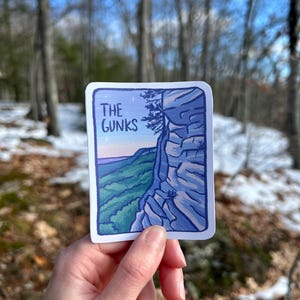 May include: A rectangular sticker featuring a landscape illustration of a mountain range. The sticker has a blue border and the text "THE GUNKS" in white. The illustration depicts a cliff face and a valley with green and purple hues.