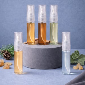 May include: Five small, clear spray bottles containing liquids of different colours, including amber, green, and blue, are arranged on a grey platform against a blue backdrop. Decorative elements such as pine needles and crystals are also present.