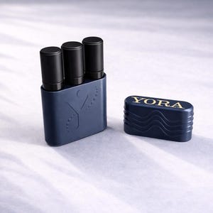 May include: A navy blue case containing three black cylindrical objects. Beside it is a matching navy blue rectangular container with gold text reading "YORA". The case has a stylized design. The containers are on a white surface.