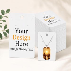 May include: A white rectangular jewellery display card with the black and orange text Your Design Here image/logo/text, alongside a stack of identical cards and a silver dog tag necklace featuring a sunset silhouette of people on a beach.