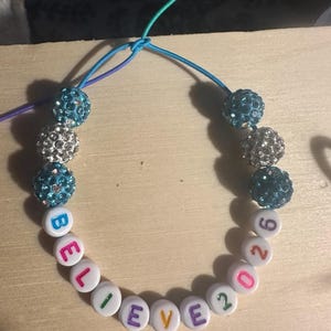 Puede incluir: Una pulsera hecha a mano con cuentas de letras blancas que deletrean "BELIEVE 2026", intercaladas con cuentas azules y plateadas cubiertas de pedrería. La pulsera está ensartada en un cordón colorido, con cierre de nudo.