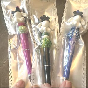 May include: Three pens individually packaged in clear plastic sleeves. Each pen features a white rabbit-like character topper with black ears, a textured bead accent in green, lime, or blue, and a solid barrel in pink, black, or blue.