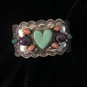 May include: A silver cuff bracelet with a heart-shaped turquoise stone in the center. The bracelet is adorned with oval-shaped coral and amethyst stones, and a scalloped edge design. The bracelet is approximately 2 inches wide.