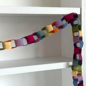 Puede incluir: Una guirnalda de cadena de fieltro de colores con eslabones en varios colores, incluyendo rosa, morado, azul, verde, naranja y amarillo. La guirnalda está colocada sobre una estantería blanca, creando un acento decorativo. La cadena es de fieltro.