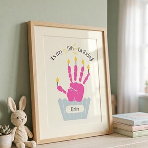 Puede incluir: Un cuadro enmarcado con una huella de mano rosa que se asemeja a una magdalena con cinco velas amarillas. El texto dice "It's my 5th birthday!" y "Erin" está escrito en el molde de la magdalena. El marco es de madera clara.