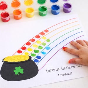 Puede incluir: Un proyecto de arte con temática del Día de San Patricio con un arcoíris hecho de huellas dactilares y una olla de oro. Las palabras "Lucky Us... We Found the Rainbow!" están impresas en el papel. Los botes de pintura de varios colores están colocados arriba.