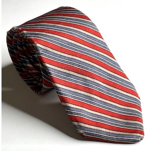 Puede incluir: Una corbata con un patrón repetitivo de rayas horizontales en rojo vibrante, azul apagado y blanco roto. La tela muestra una textura fina y la corbata está doblada sobre sí misma, mostrando el extremo puntiagudo y la construcción en capas.