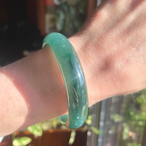 May include: A smooth, polished, translucent green bangle bracelet with subtle darker green inclusions and a rounded profile. The material has a glossy, reflective surface that catches the light, showcasing a deep, vibrant emerald green hue throughout.