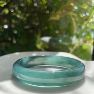 Puede incluir: Un brazalete rígido liso y pulido hecho de jadeíta verde translúcida. La banda circular presenta un perfil redondeado con un acabado brillante, mostrando variaciones naturales en la intensidad del color e inclusiones blancas nubladas.