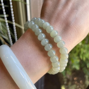 May include: A smooth, solid white bangle bracelet and two beaded bracelets made of polished, pale green, translucent spherical beads. The jewelry pieces are worn together, showcasing the contrast between the opaque white band and the light green beads.