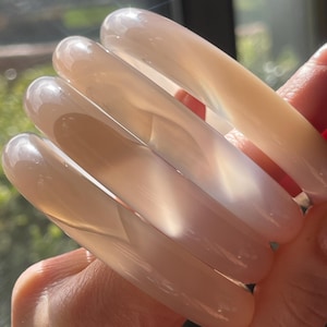 May include: A stack of three smooth, translucent, pale beige bangle bracelets. The circular bangles have a polished, glossy finish that catches the light, revealing subtle internal variations in the light tan and off-white material.
