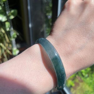 May include: A smooth, polished bangle bracelet featuring a deep forest green color with subtle, translucent variations and cloudy white inclusions throughout the solid, rounded band, worn around the wrist to showcase its natural stone-like appearance.