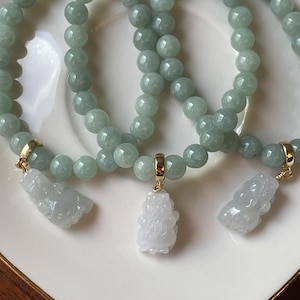 May include: Three beaded bracelets made of smooth, light green jade spheres. Each bracelet features a gold-toned metal bail holding a carved, translucent white jade pendant shaped like a mythical creature, suspended from the circular beaded strand.