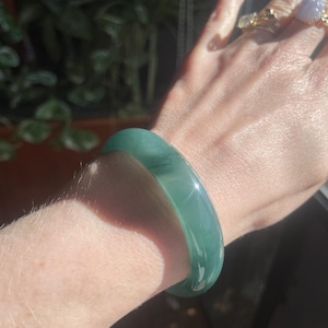 May include: A smooth, translucent green jade bangle bracelet with a polished finish, worn on the wrist. The circular band features deep emerald green tones with subtle natural variations and light-reflecting surfaces.