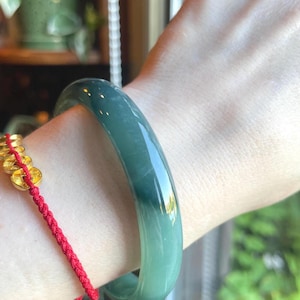 May include: A smooth, polished, translucent dark green jade bangle bracelet with a rounded profile. The bracelet features subtle variations in color intensity and is worn alongside a thin red cord bracelet adorned with small gold-colored metal coins.