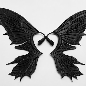 May include: A pair of matte black ear cuffs shaped like intricate butterfly wings. Each cuff features a curved structure that hooks around the ear, with detailed vein patterns and pointed edges extending outward to mimic the appearance of insect wings.
