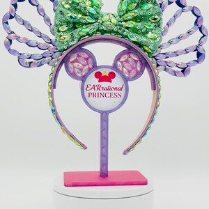 May include: A decorative headband with a green sequined bow and purple accents. The headband features a Minnie Mouse ear design with the text "EARrational PRINCESS". The ears and headband are adorned with iridescent gems.