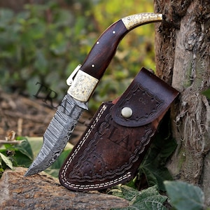 Könnte beinhalten: Ein Klappmesser mit einer Damaststahlklinge, einem dunklen Holzgriff und Messingdetails. Es wird von einer braunen Lederscheide mit dekorativen Nähten und einem Druckknopf begleitet. Die Klinge weist ein unverwechselbares Muster auf.