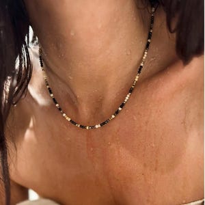 Puede incluir: Un delicado collar compuesto por pequeñas cuentas redondas en tonos negro, blanco y dorado. El collar se lleva alrededor del cuello, mostrando las sutiles variaciones de color y el intrincado diseño. Las cuentas parecen estar ensartadas, creando un accesorio refinado.