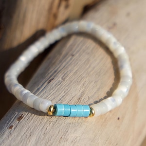 May include: A white beaded bracelet with a turquoise accent. The bracelet features small, round white beads and a central section of turquoise beads, separated by gold-colored accents. The bracelet is displayed on a wooden surface.