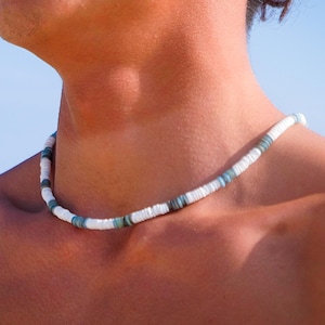 Puede incluir: Un collar de cuentas blancas y turquesas. El collar está compuesto por pequeñas cuentas blancas redondas intercaladas con cuentas turquesas. El collar se lleva alrededor del cuello.