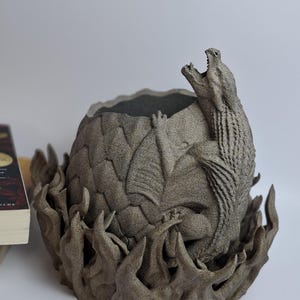 May include: A detailed, gray dragon egg-shaped planter with a dragon emerging from the top. The planter sits on a base of stylized flames. The dragon has its mouth open, and the egg has a textured surface.