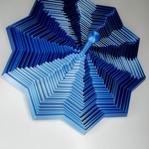 May include: A blue, star-shaped, 3D-printed object with layered, geometric design. The object has a gradient of blue hues, from light to dark, and a central handle-like structure. The design is intricate and visually striking.