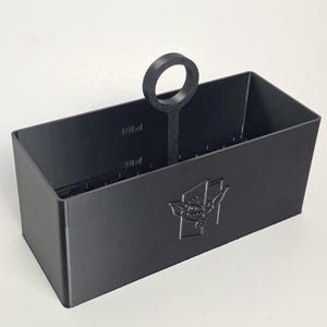 May include: A matte black rectangular storage container with a central circular handle. The front features an embossed graphic of a creature peeking through a doorway. The interior walls display printed measurement markings up to 3000 ml.