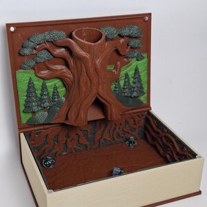 May include: A book-shaped dice tower and tray featuring a brown 3D-printed tree trunk with a hollow top for dice insertion. The interior back panel displays a green landscape with dark green pine trees. The base is brown with root details.