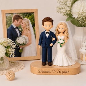Pode incluir: Um topo de bolo de casamento com duas figuras numa base de madeira clara gravada com o texto Brooks ♡ Skylar. A figura masculina veste um fato azul-marinho com laço preto, e a feminina usa um vestido de renda branco e um véu.
