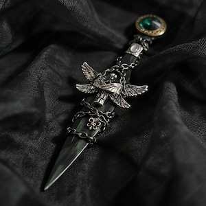 Puede incluir: Una daga ornamentada con una hoja verde oscuro y detalles plateados intrincados. El mango presenta una gema verde, alas de ángel y una cadena. La daga se encuentra sobre un fondo oscuro y texturizado. El texto "HearthstoneCraftUS" es visible.