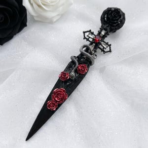 Puede incluir: Una daga negra con adornos de rosas rojas y serpientes plateadas. El mango presenta una rosa negra y una cruz con una gema roja. La hoja está decorada con rosas rojas.