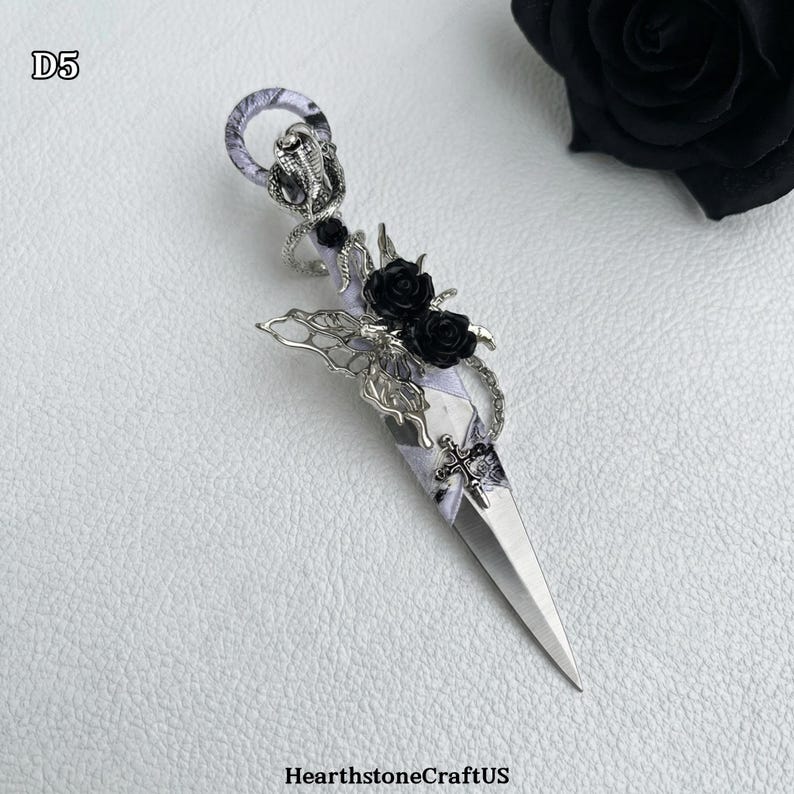 May include: A decorative dagger with a silver blade and ornate handle. The handle features a snake, black roses, and a cross design. The dagger is embellished with silver filigree and a patterned fabric wrap. The text "D5" is in the upper left corner.