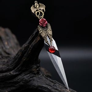 May include: A decorative dagger with a silver blade and ornate bronze accents. The handle features a red rose, a red gem, and a bronze dragon and wing design. The dagger is displayed against a dark, textured background.